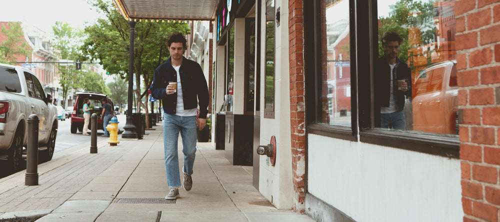 A person walks down a sidewalk holding a drink in an urban area with parked cars and storefronts.