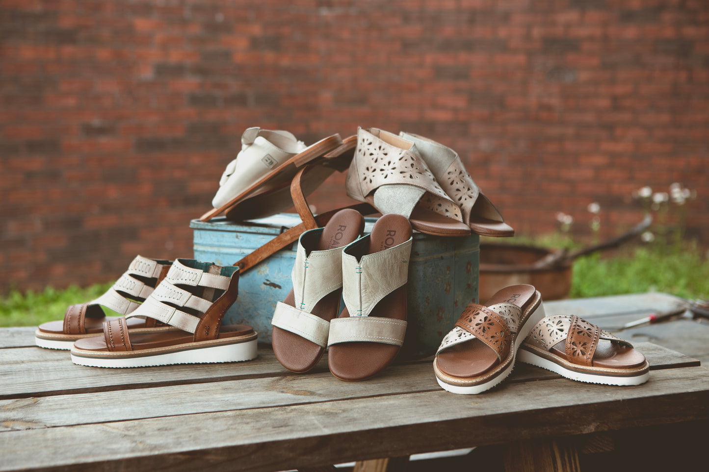 A collection of sandals with various designs and colors are arranged on a rustic wooden table, with a brick wall and greenery in the background.