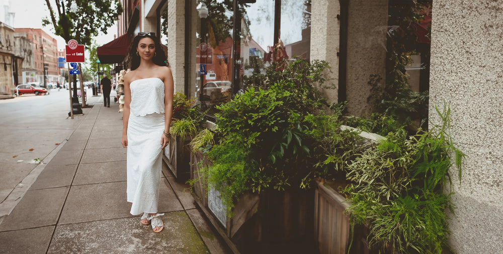 A woman in a long, strapless white dress walks down a city sidewalk beside a row of green plants in planters. She has long hair and is wearing sandals.