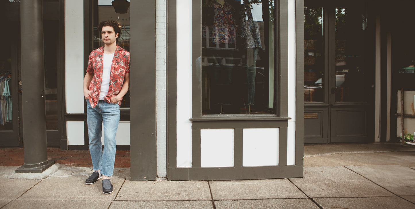 A person stands on the sidewalk in front of a storefront, wearing a red patterned shirt, white t-shirt, blue jeans, and dark shoes.