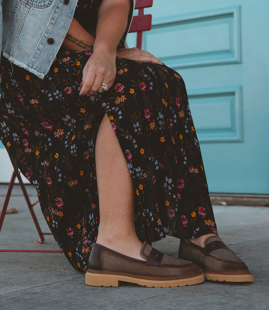 A person sits on a red chair, dressed in a denim jacket and floral dress, elegantly accessorized with Roan loafers in brown from the Beginning collection, showcasing the luxurious experience of women's footwear. One hand rests on their knee against a blue door backdrop.