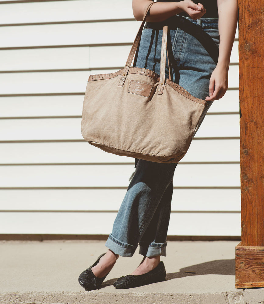 
                  
                    A person wearing Roan jeans and Roan slip-on ballerina shoes, holding a beige Business tote bag, leans against a wooden post in front of a white siding wall.
                  
                