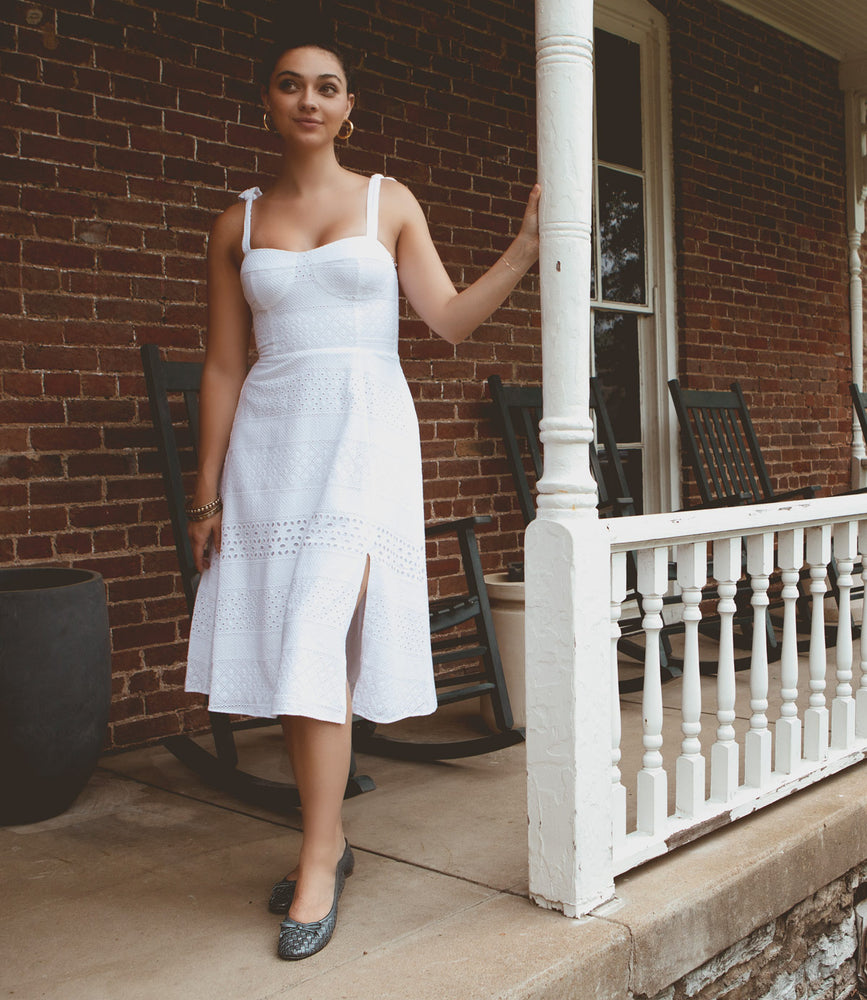 
                  
                    A woman in a white dress stands on the porch of a brick building, holding a white porch column. There are rocking chairs behind her, showcasing the intentional craftsmanship of the hand-woven leather cushions that invite relaxation, featuring Roan's Business model.
                  
                