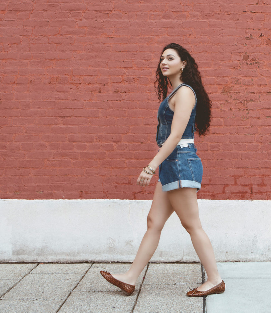 
                  
                    A person with long curly hair wearing a blue sleeveless top, shorts, and Roan Business slip-on ballerina shoes walks on a sidewalk in front of a red brick wall.
                  
                