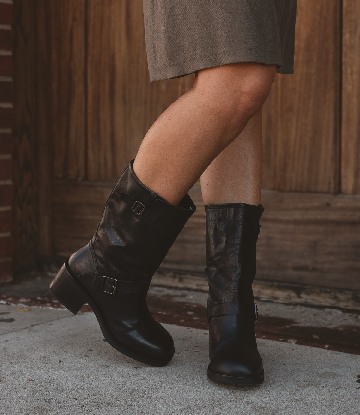 
                  
                    A person wearing the Roan Cite black leather mid-calf boots with buckles and a contemporary edge stands on a sidewalk, with a wooden door in the background.
                  
                