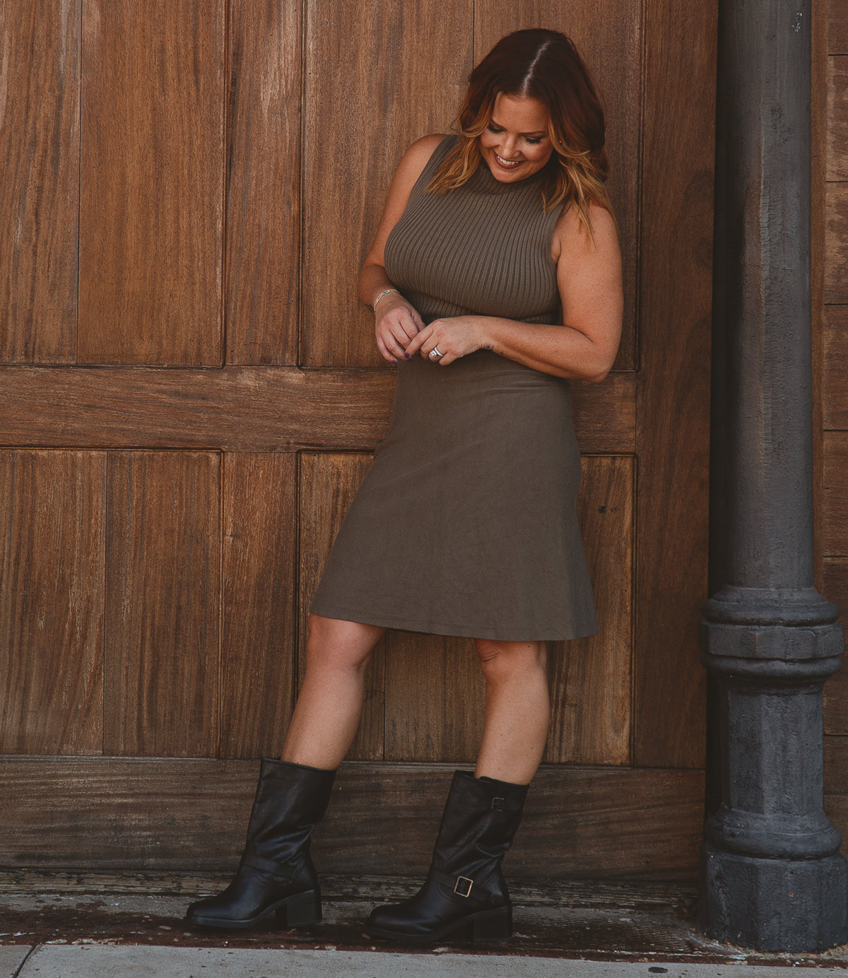 
                  
                    A person wearing the sleeveless "Cite" dress by Roan, paired with modern boots, stands against a wooden wall, glancing down with a subtle smile.
                  
                