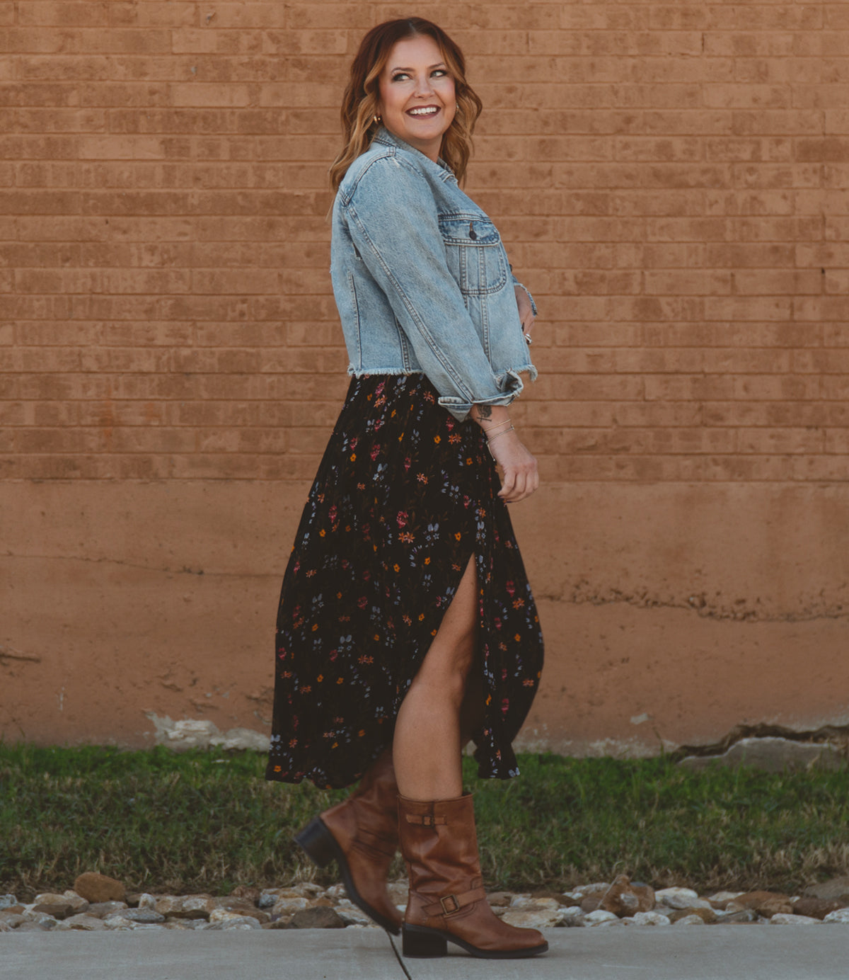 
                  
                    A modern woman in a denim jacket and floral dress strolls along the sidewalk, smiling. Her Roan Cite brown boots with a square toe design perfectly complement her casual autumn outfit. The background features a red brick wall and green grass, adding charm to her stylish ensemble.
                  
                