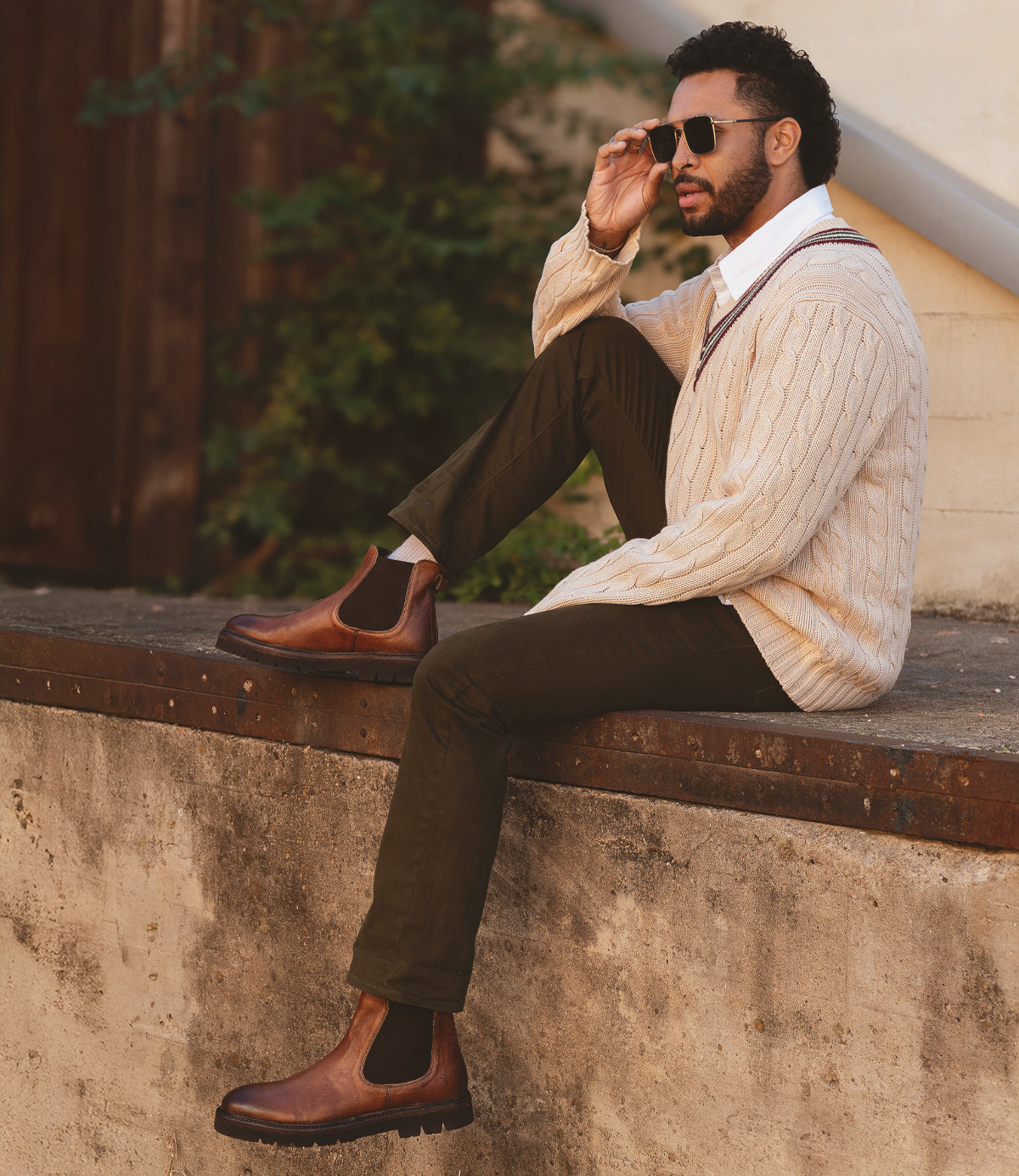 
                  
                    A man sits on a concrete ledge, wearing Roan's Defy sunglasses, a cream cable-knit sweater, dark pants, and brown Chelsea boots with a traction-friendly sole. He gazes into the distance with one hand adjusting his Defy sunglasses.
                  
                