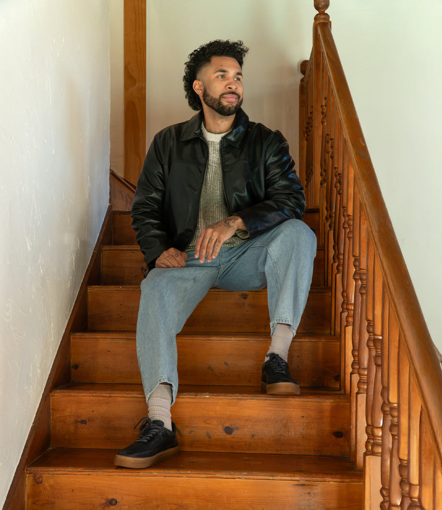 
                  
                    A person with curly hair sits on wooden stairs, wearing a black jacket, gray sweater, and blue jeans. They gaze off to the side, their feet comfortably supported by Roan's durable "Eminent" leather sneakers.
                  
                