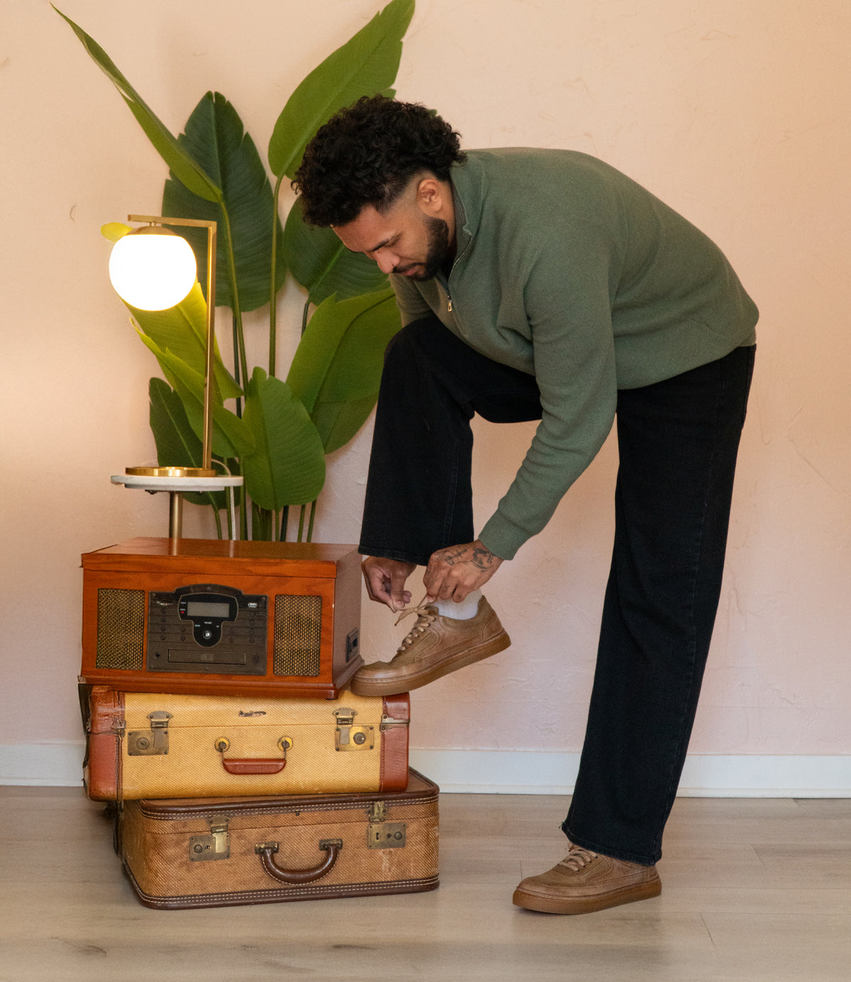 
                  
                    A person in a green sweater ties their Roan Eminent footwear, standing beside stacked vintage suitcases and a retro radio under a lamp and plant.
                  
                