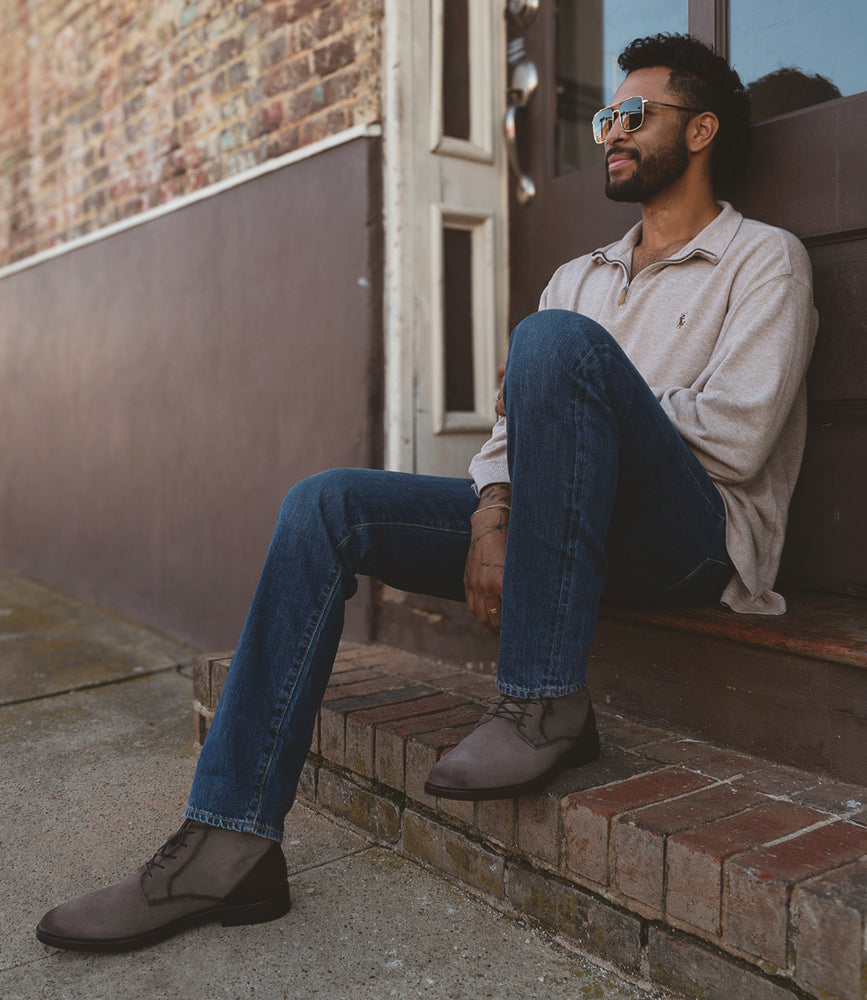 
                  
                    A man in sunglasses sits on a brick step outside, wearing a light sweater, jeans, and gray suede shoes called Joyride by Roan. The brick and wood wall behind him perfectly complements his laid-back style.
                  
                