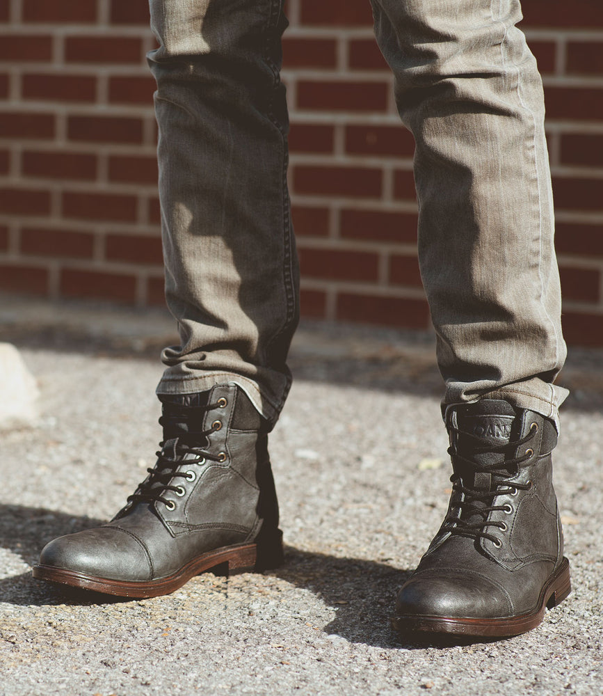 
                  
                    A person wearing Roan's Lecture, full-grain leather lace-up boots in gray with a padded collar, paired with faded jeans, stands on a gravel surface in front of a brick wall.
                  
                