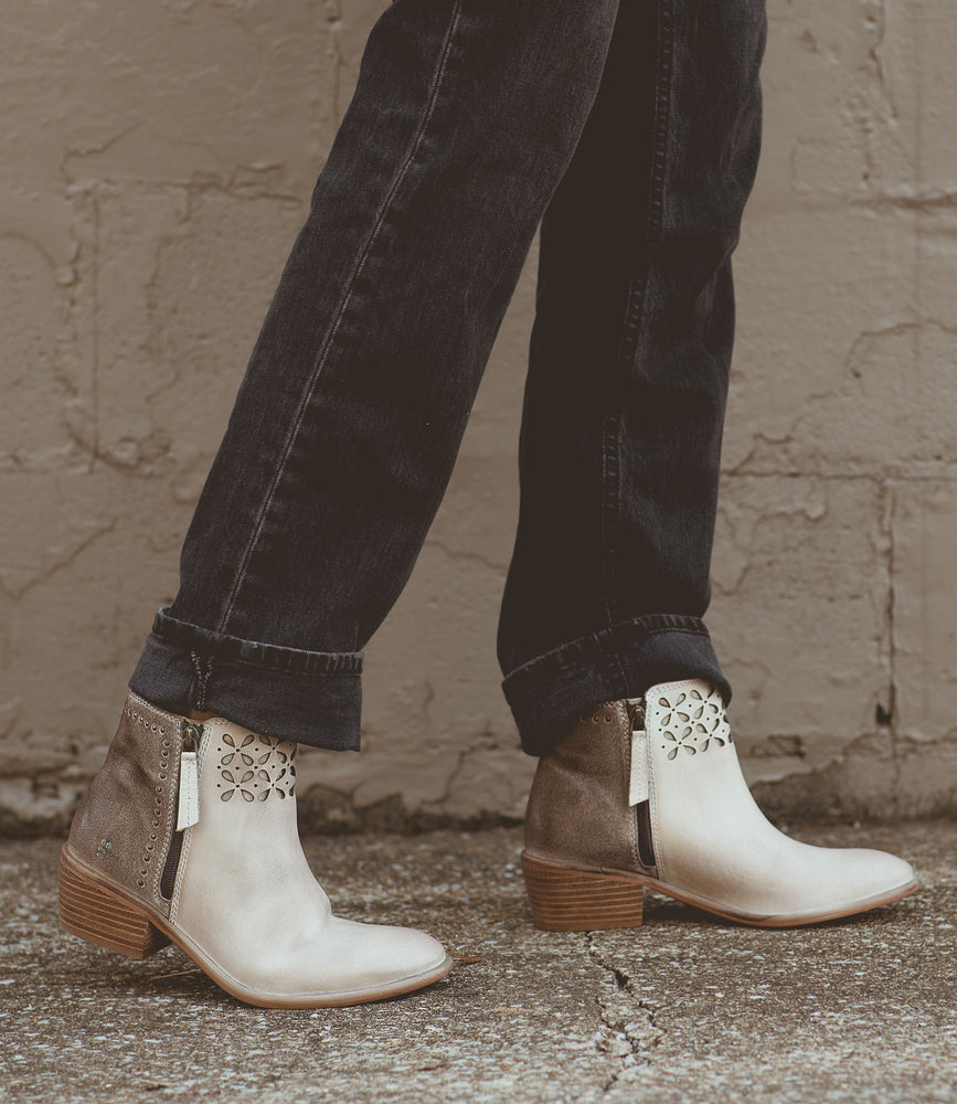 A person wearing the Roan Liz Cutout two-tone ankle boots with a petal design, paired with black jeans, standing on a textured surface.