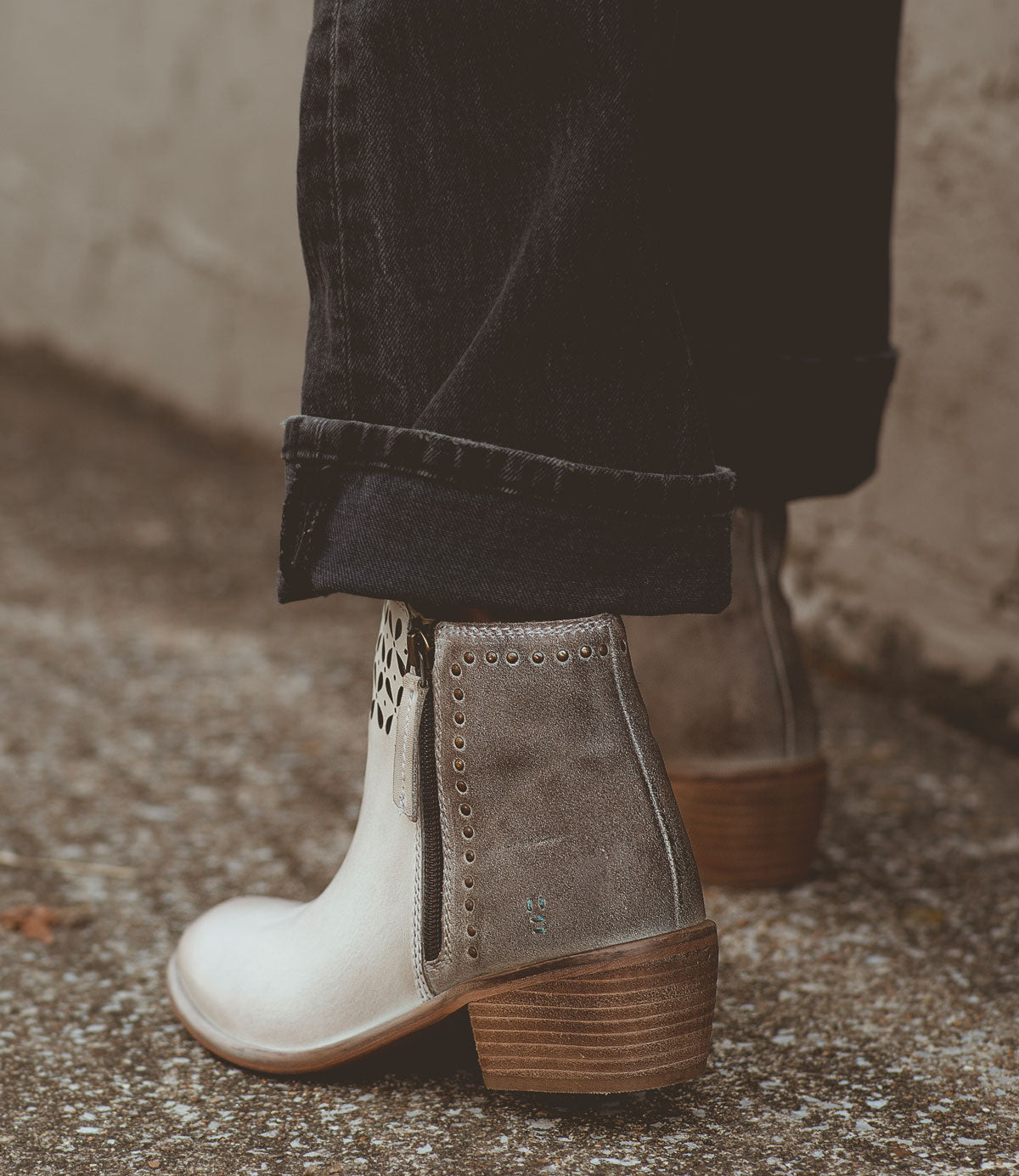 
                  
                    A person wearing black pants and the stylish Roan Liz Cutout ankle boots, featuring gold rivet details and a wooden heel, stands on a textured pavement.
                  
                