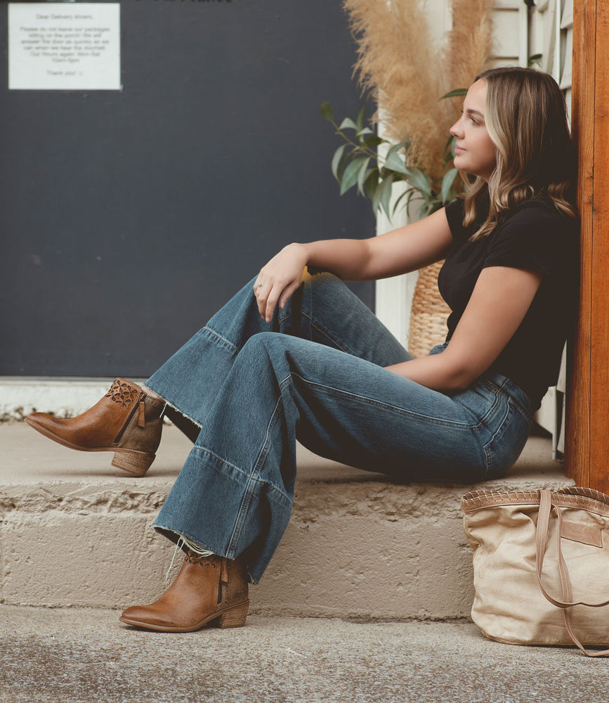 
                  
                    A woman is sitting on a step, wearing the Roan Liz Cutout black top paired with wide-legged jeans and brown ankle boots, with a beige bag beside her.
                  
                