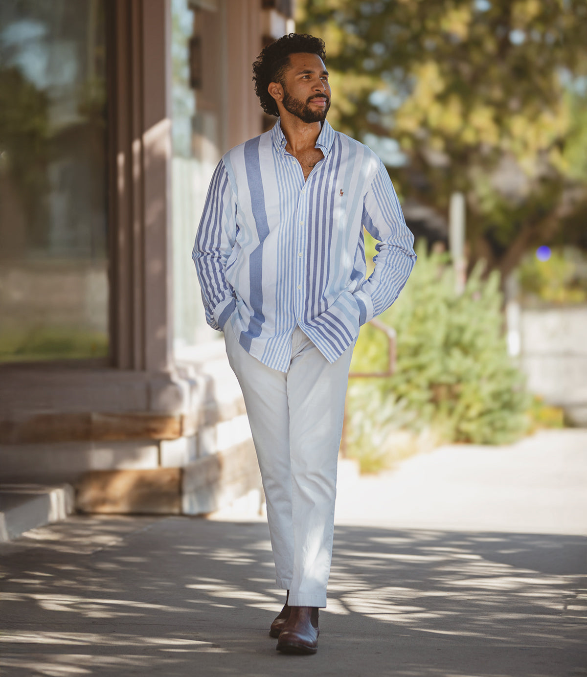 
                  
                    A man strolling outside wears Roan's Looting shirt in light blue and white stripes, paired effortlessly with white pants and brown Chelsea boots crafted from full-grain leather, embodying both comfort and style.
                  
                