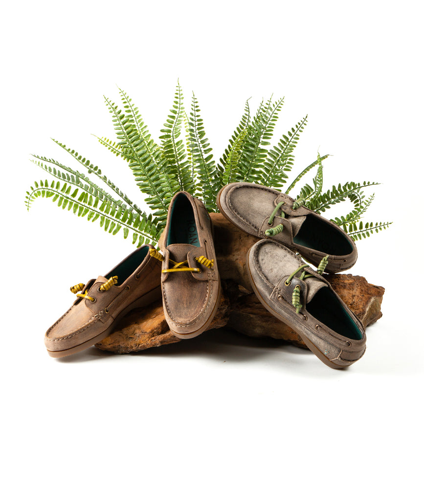 Three tan leather Abide boat shoes by Roan, featuring brown and green shoelaces, are displayed on a wooden surface surrounded by green ferns.