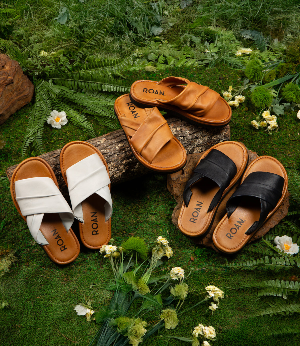 Three pairs of Roan Nosy slip-on sandals, in white, brown, and black with leather footbeds and "ROAN" inscribed on the insoles rest on grass and logs. Green ferns and white flowers enhance the natural charm of the scene.