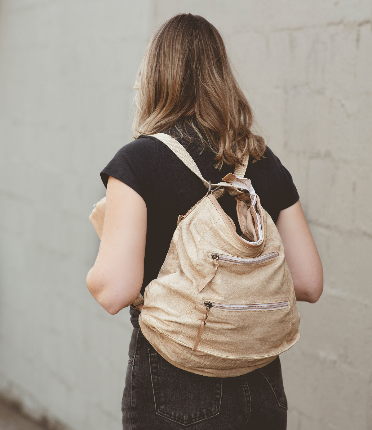 
                  
                    A person wearing a black shirt and jeans is carrying the Roan Oganness, a versatile beige backpack perfect for any travel companion. They stand confidently against a gray wall.
                  
                