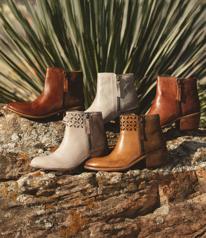 Five Roan Liz Cutout ankle boots in brown and gray sit on rocks, each adorned with gold rivets. The elegant design is enhanced by a plant gracefully framing the background.