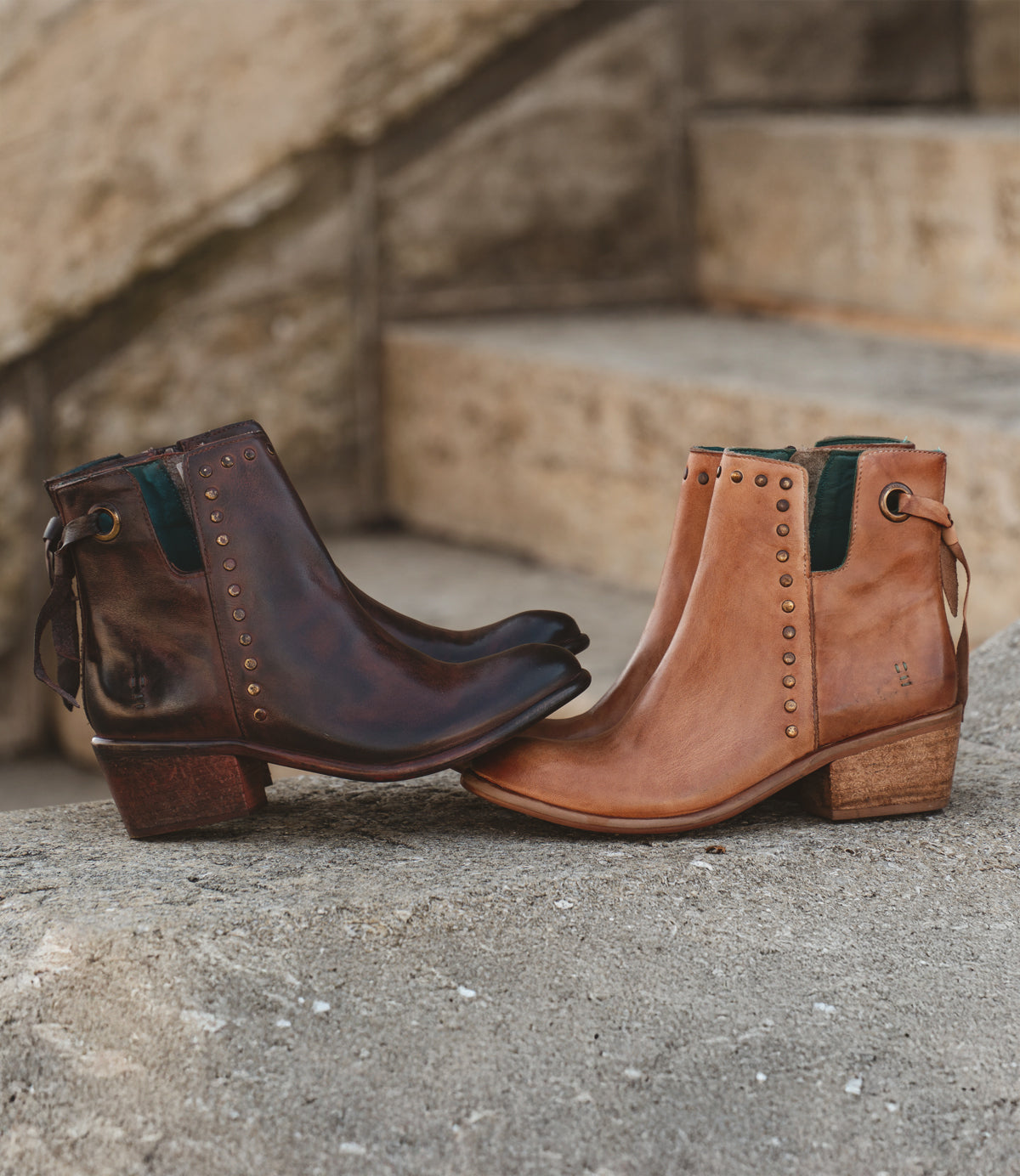 Placed on stone steps, Roan's Australia boots embrace the western trend with one dark brown and the other tan. These leather boots feature rounded toes, low heels, metal studs, and pull tabs.