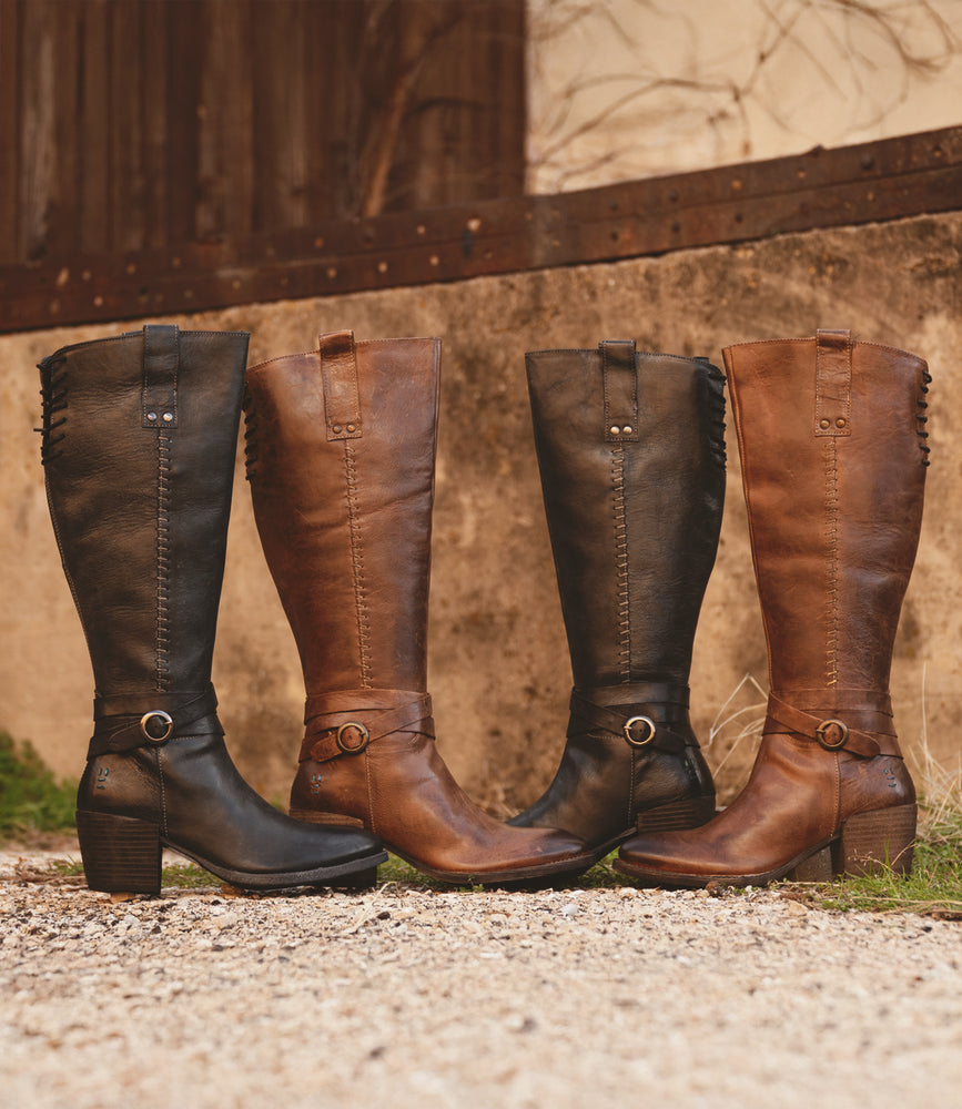 Four Garner knee-high leather boots from Roan, featuring a chic ankle strap and moderate heel, stand upright on a gravel surface against a rustic wall. These black and brown beauties exude timeless style.