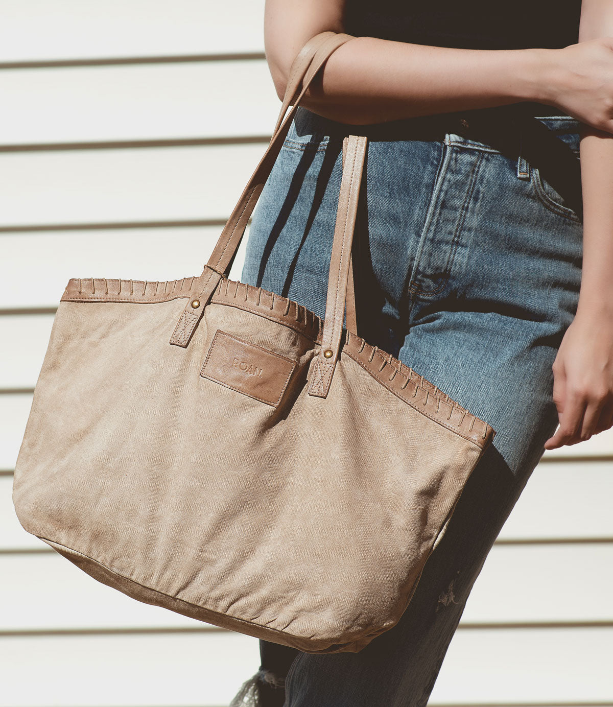 
                  
                    A person holding the Rutherford, a large beige tote bag from Roan, made of lightweight cotton canvas with long handles and a small label on the front, standing against a striped wall. This handmade tote exudes timeless style.
                  
                