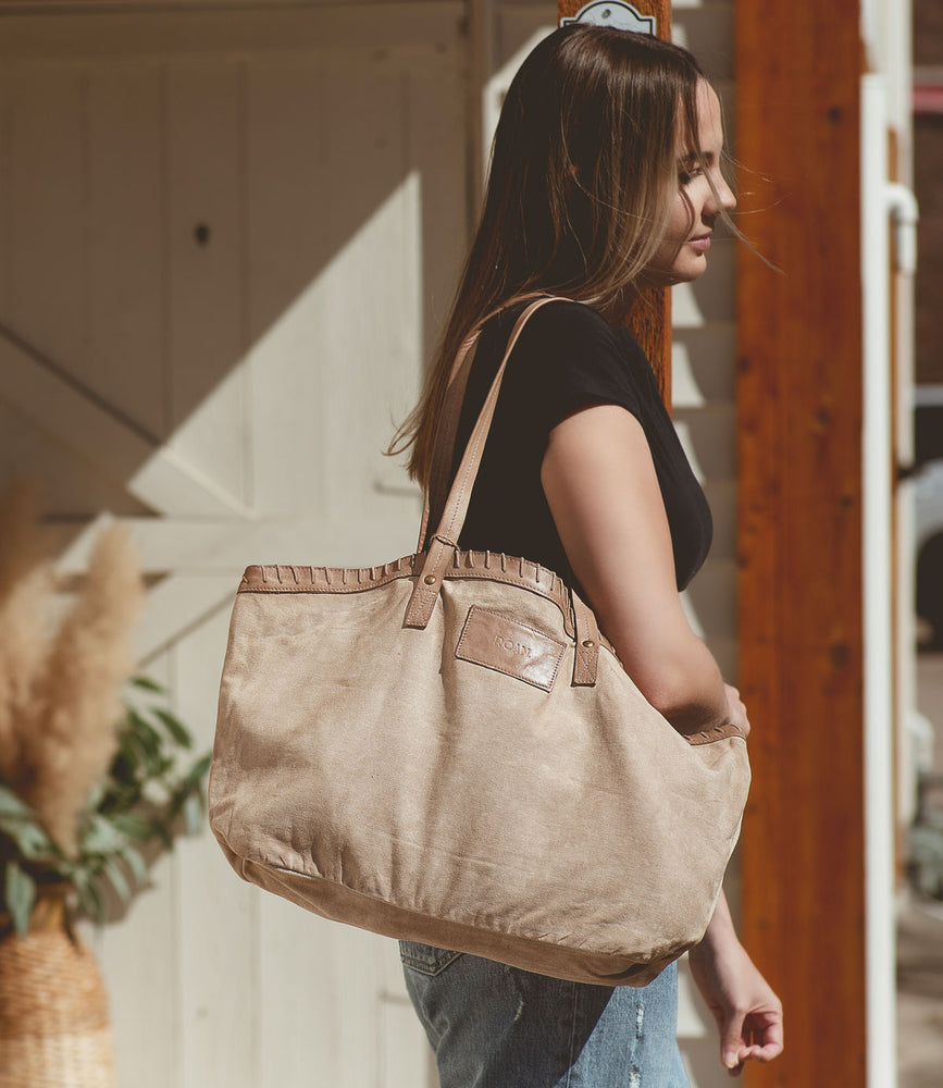 
                  
                    A woman with long hair carries a Roan Rutherford tote, made from lightweight cotton canvas, over her shoulder while standing in front of a wooden door, exuding timeless style.
                  
                