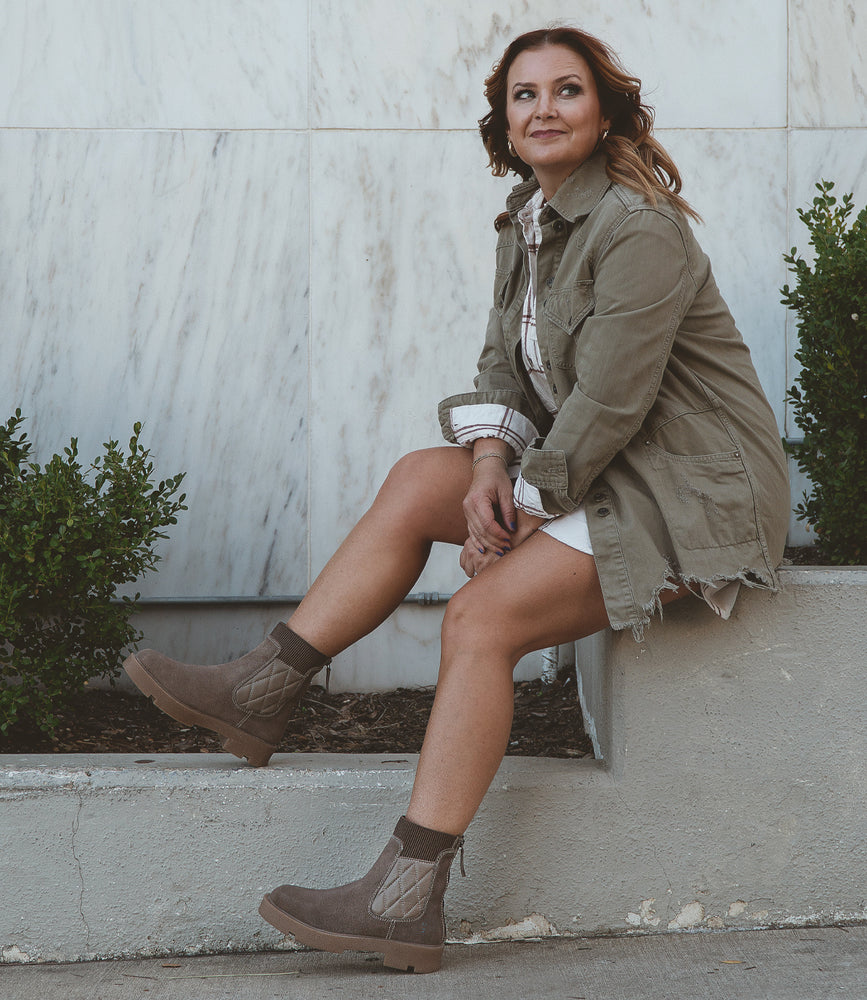 
                  
                    Wearing the Roan Thoughtful jacket and stylish boots, a person relaxes on a concrete ledge near green shrubs, embodying an effortless blend of comfort and style against the marble wall backdrop.
                  
                