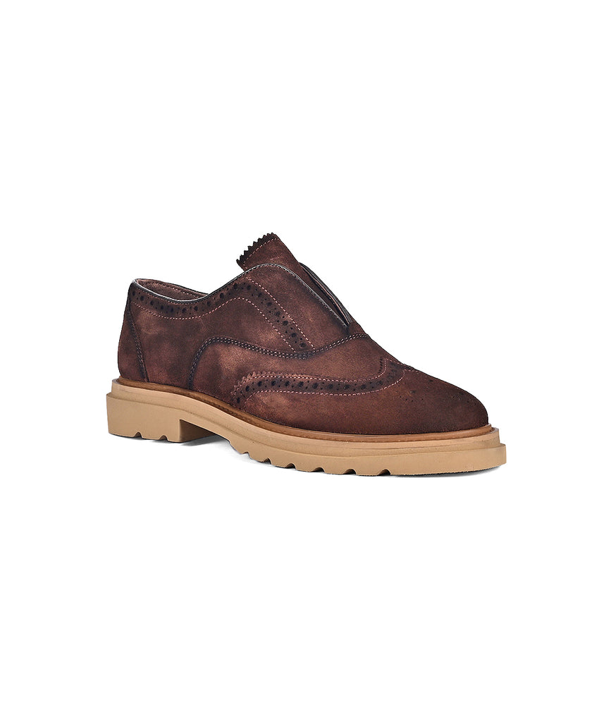 The "Blame" shoe by Roan is a brown suede slip-on adorned with brogue detailing and set on a chunky beige rubber sole.